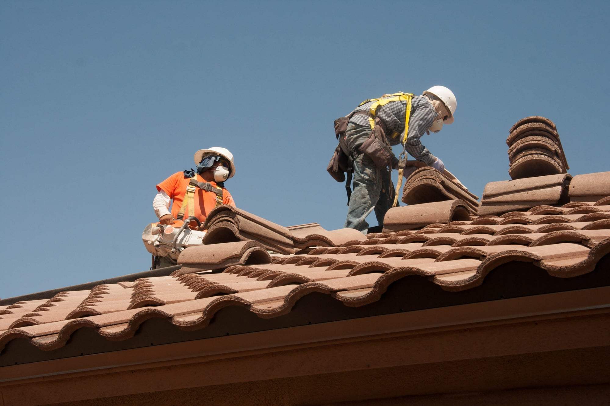 Two roofers in safety harnesses resetting clay barrel tiles along the edge of a roof.