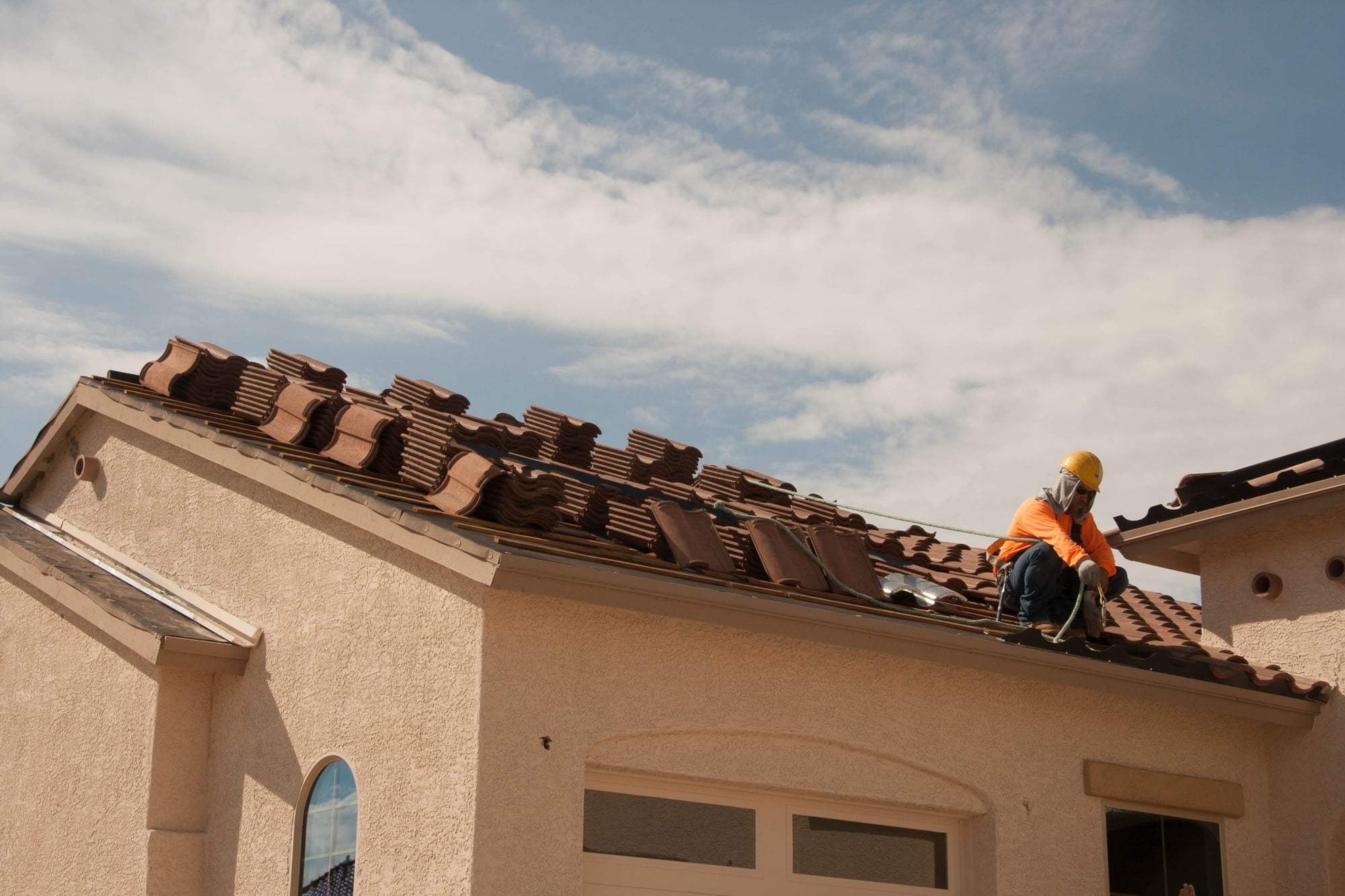 A roofer working across the tile roof of a stucco home under a wide sky.